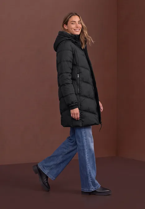 Lange Pufferjacke mit Kapuze Black Frau in schwarzem Steppmantel, blauen Jeans und schwarzen Schuhen posiert seitlich vor braunem Hintergrund.