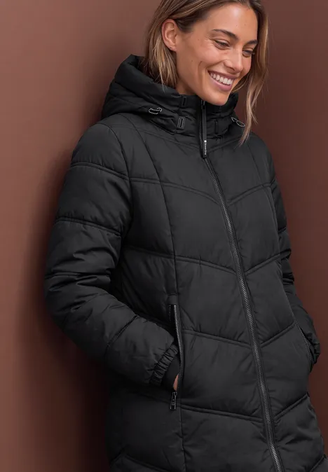 Lange Pufferjacke mit Kapuze Black Frau lächelt in schwarzer, wattierter Kapuzenjacke vor braunem Hintergrund.