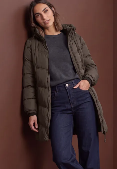 Lange Pufferjacke mit Kapuze auburn brown Frau in olivgrünem Daunenmantel, dunklem Pullover und Jeans vor braunem Hintergrund.
