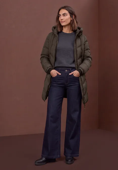 Lange Pufferjacke mit Kapuze auburn brown Eine Frau in dunkler Winterjacke, grauem Pullover und weiten Jeans steht vor braunem Hintergrund.