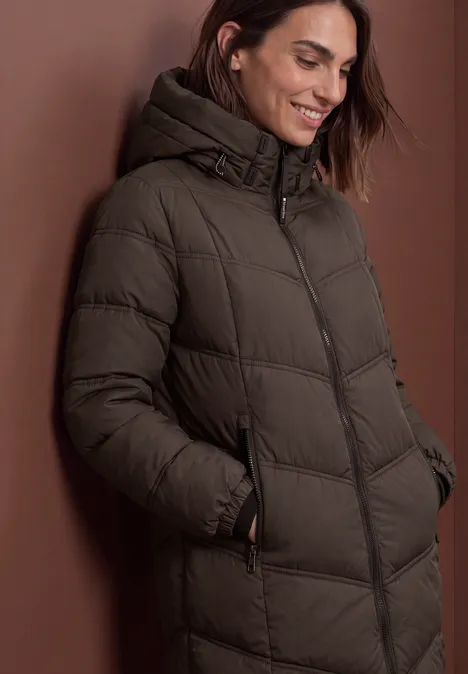Lange Pufferjacke mit Kapuze auburn brown Frau in olivgrünem, gesteppter Wintermantel mit Kapuze lehnt vor braunem Hintergrund.