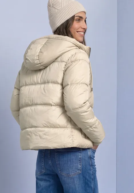 Frau trägt helle, wattierte Daunenjacke mit Kapuze und Beanie, sieht über ihre Schulter und lächelt.