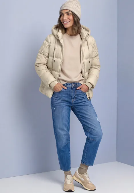 Frau in beige Steppjacke, Beanie, blauen Jeans und Sneakers vor schlichtem Hintergrund.