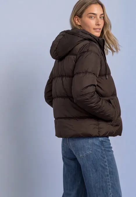 Eine Frau in brauner Steppjacke mit Kapuze und blauen Jeans vor hellem Hintergrund.
