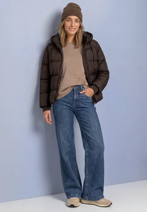 Frau steht lächelnd in dunkler Steppjacke, hellem Pullover, Jeans und braunen Boots vor neutralem Hintergrund.