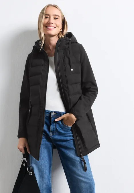 Softshelmantel mit Kapuze und Steppdetails Black Frau trägt schwarzen Parka mit Kapuze, weiße Bluse und blaue Jeans, schaut lächelnd in die Kamera.