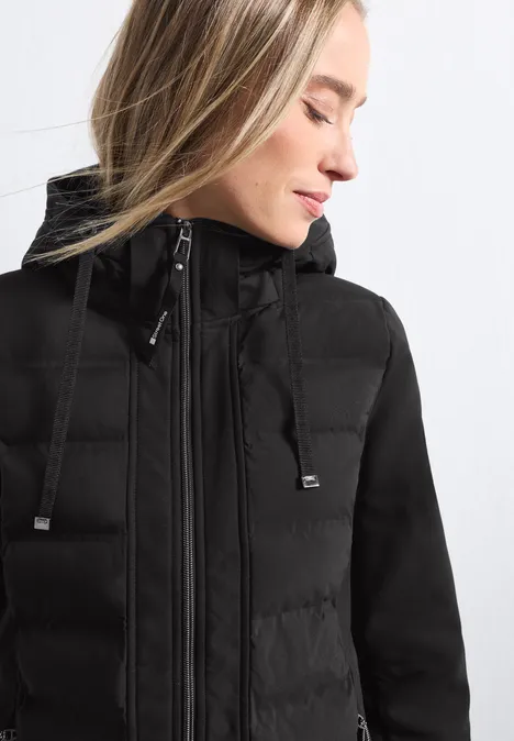 Softshelmantel mit Kapuze und Steppdetails Black Frau mit schwarzer Steppjacke, hohem Kragen und Kapuze, seitliche Nahaufnahme.