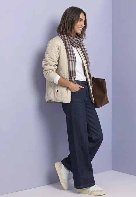 Blouson met doorgestikt patroon en ritssluiting barley beige Een glimlachende vrouw voor een paarse muur, met een beige jack, geruite sjaal, wijde jeans, witte sneakers en bruine tas.