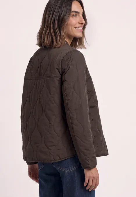 Frau in brauner, gesteppter Jacke, seitlich stehend und zurückblickend, kombiniert mit Jeans.