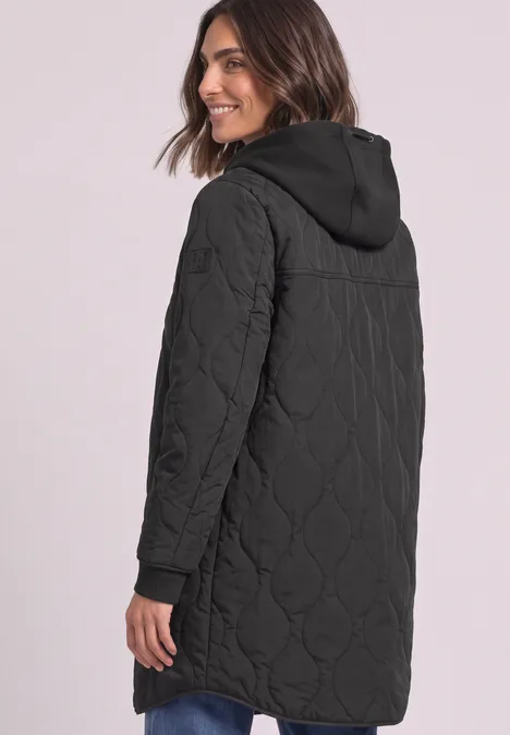 Eine Frau trägt eine schwarze, gesteppte, knielange Jacke mit Kapuze und lächelt seitlich.