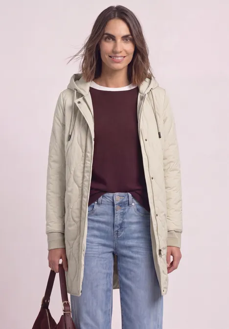 Eine Frau trägt einen hellen Parka, ein bordeauxfarbenes Oberteil und Jeans, und hält eine Handtasche.