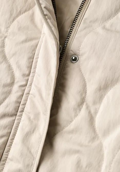 Nahaufnahme einer hellbeigen Steppjacke mit sichtbarem Reißverschluss und Druckknopf.