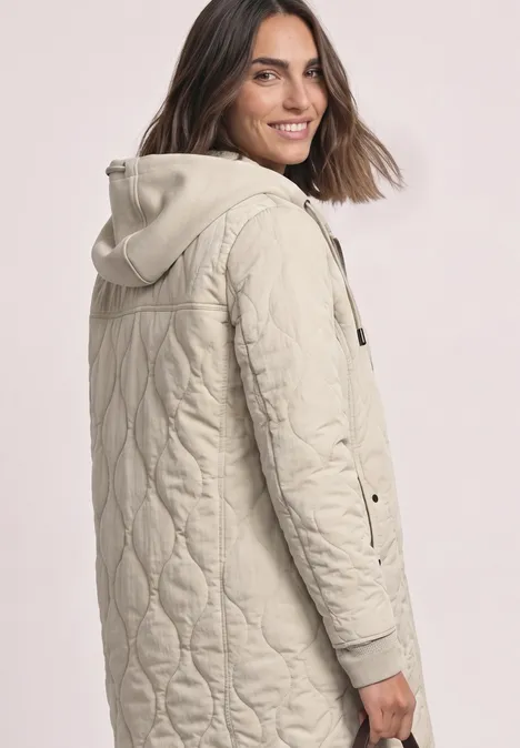 Eine lächelnde Frau trägt eine hellbeige, gesteppte Kapuzenjacke mit seitlichen Druckknöpfen.