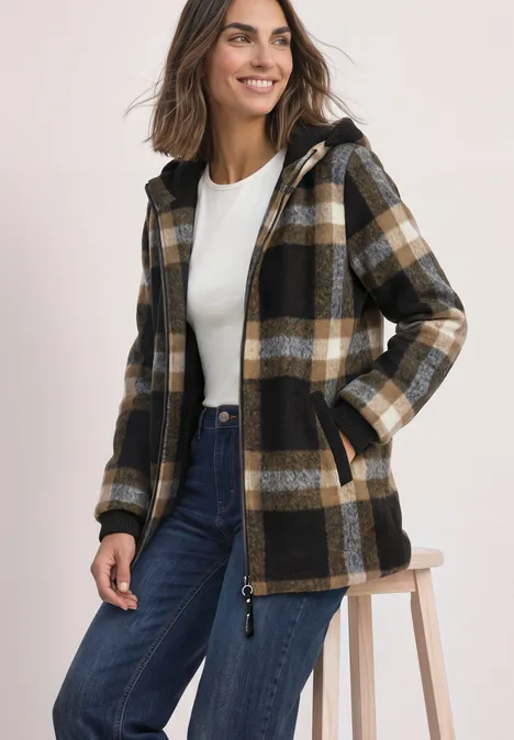 Frau trägt karierte Kapuzenjacke, weißes Top und Jeans, sitzt auf Hocker und lächelt.