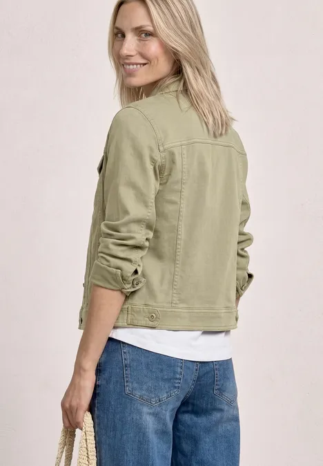Von hinten: Frau mit blonden Haaren trägt olivgrüne Jeansjacke, weißes Shirt und blaue Jeans.