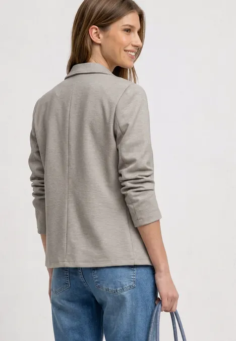 Een persoon gezien van achteren draagt een lichtgrijs jasje, blauwe jeans en houdt een tas vast.