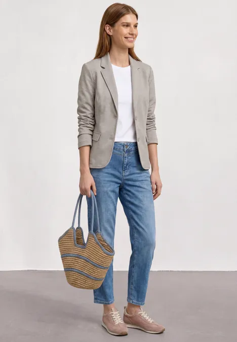 Vrouw draagt spijkerbroek, wit shirt, grijs jasje en strooien tas.