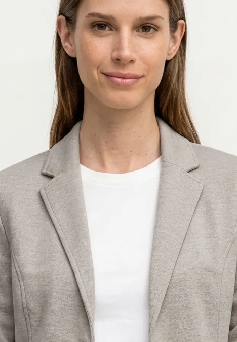 Bovenlichaam van een persoon in beige colbert en wit T-shirt, gezicht vervaagd.