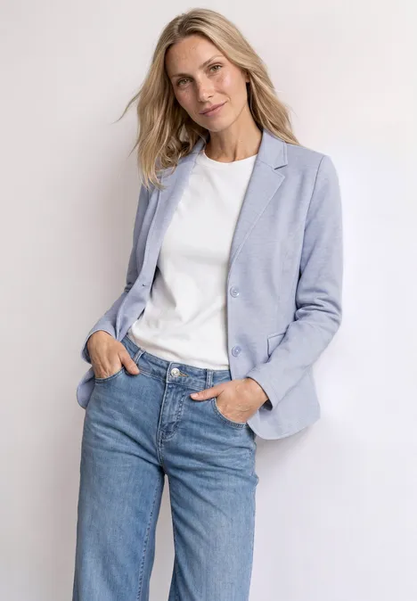 Basic Piqué Blazer whispery blue Eine Person trägt einen hellblauen Blazer, weißes Shirt und blaue Jeans, steht lässig mit Händen in den Taschen.