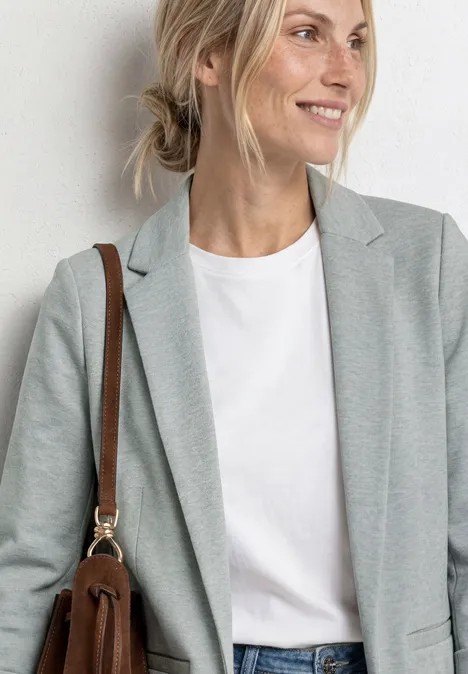 Blazer im langen und offenen Schnitt bound green Frau in grauem Blazer, weißem T-Shirt und brauner Schultertasche vor neutralem Hintergrund.