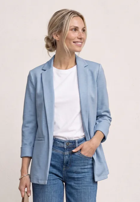 Femme en blazer bleu clair, t-shirt blanc et jean, pose décontractée avec une main dans la poche.