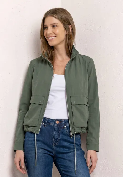 Frau in grüner Jacke, weißem Shirt und blauer Jeans steht vor hellem Hintergrund.