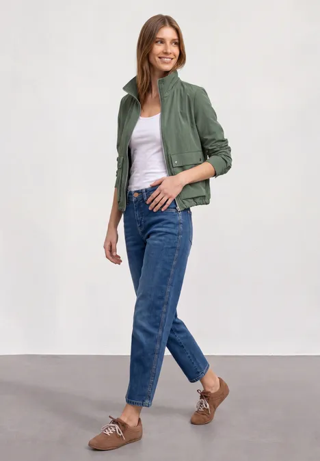 Eine Frau trägt eine grüne Jacke, weißes Top, blaue Jeans und braune Slingback-Schuhe.
