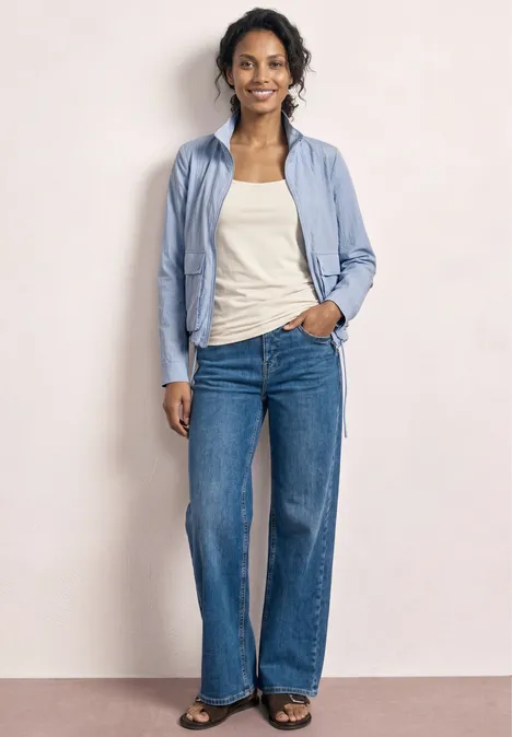 Frau vor hellem Hintergrund, hellblaue Bluse, weißes Top und weite blaue Jeans.
