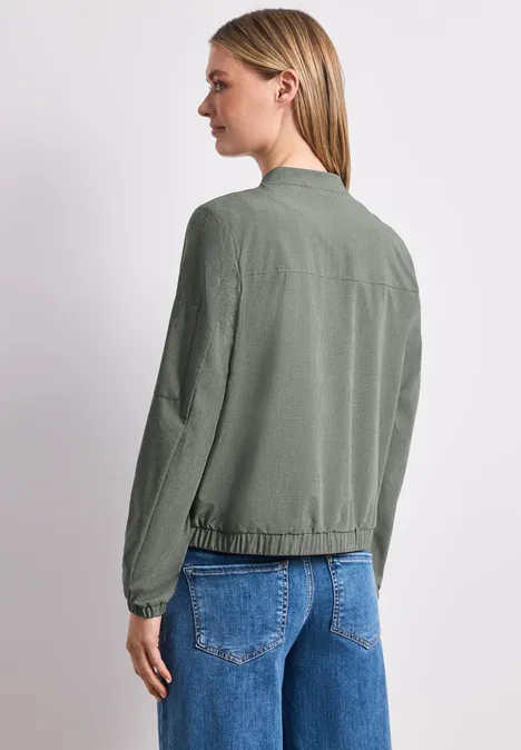 Vrouw van achteren in olijfgroene bomber en blauwe jeans voor een lichte achtergrond.