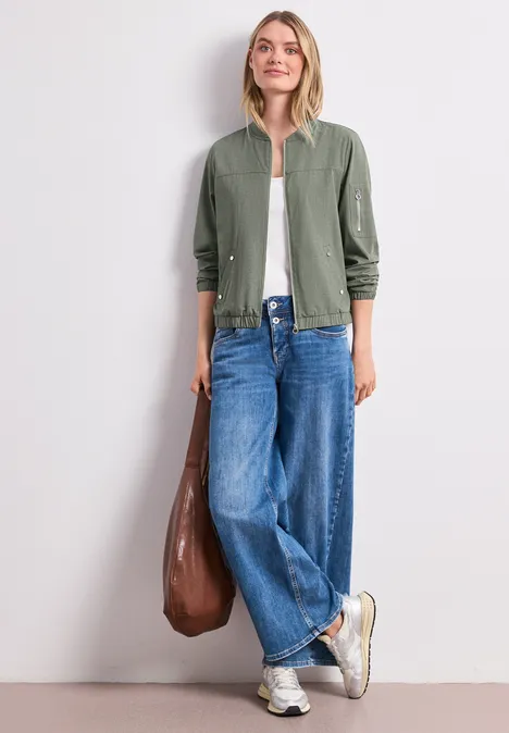 Persoon met groen jasje, wit topje, losse jeans, witte sneakers en een bruine tas voor een witte muur.