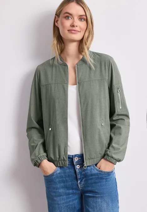 Vrouw in groene bomberjas, wit top en blauwe jeans.