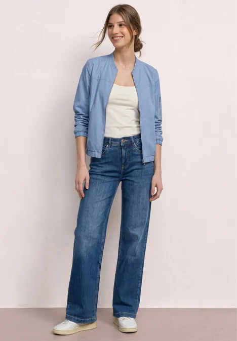 Een vrouw draagt een lichtblauw bomberjack, een wit topje, wijde high-waist jeans en witte sneakers.