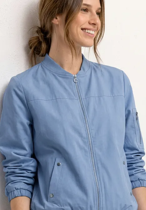Vrouw draagt lichtblauwe, getailleerde bomberjack met opstaande kraag, rits en opgestikte zakken.