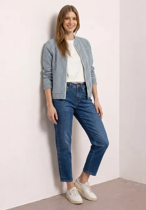 Eine Frau lehnt in heller Jacke, weißem T-Shirt, blauen Jeans und weißen Sneakern lässig an einer Wand.