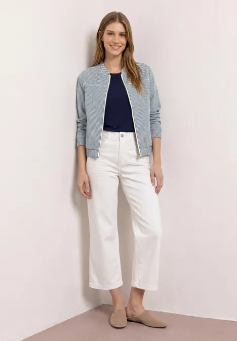 Frau in hellblauer Jacke, dunkelblauem Top, weiter weißer Jeans und beigen Slippern vor schlichtem Hintergrund.