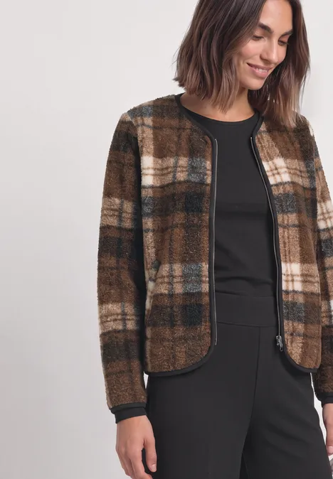 Teddy-Jacke mit Rundhals und Print Obsidian Blue Frau mit dunkelbrauner Karo-Jacke über schwarzem Oberteil und Hose lächelt.