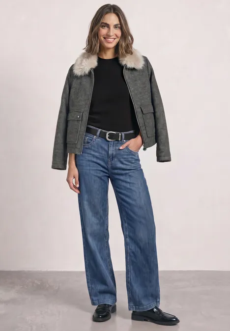 Frau in schwarzem Shirt, grüner Jacke mit Fellbesatz, weiter Jeans und schwarzen Schuhen.