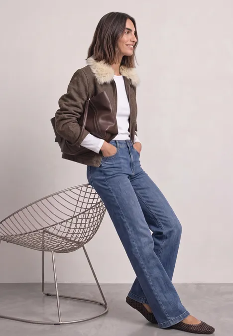 Frau in brauner Jacke mit Pelzkragen, weißem Shirt, weiten blauen Jeans und braunen Schuhen lehnt an Stuhl.
