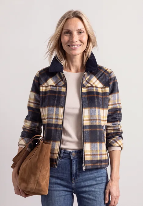 Frau mit kariertem Jacke, weißem Shirt, Jeans und brauner Tasche posiert vor neutralem Hintergrund.
