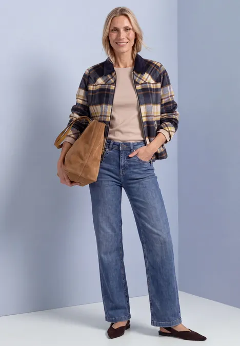 Frau in kariertem Hemd-Jacke, beigem Top und weitem Jeans, trägt braune Tasche und lächelt.