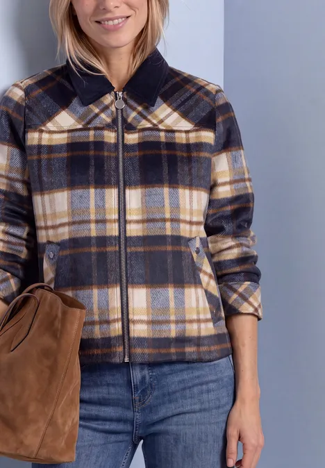 Frau trägt karierte Kurzjacke in gedeckten Farben, blaue Jeans und hält eine braune Tasche.