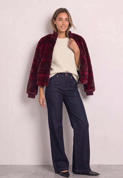 Blouson en fausse fourrure avec des détails en maille et un imprimé plummy brown Frau in bordeauxroter Kunstfelljacke, cremeweißem Pullover und weiten, dunkelblauen Jeans vor hellem Hintergrund.