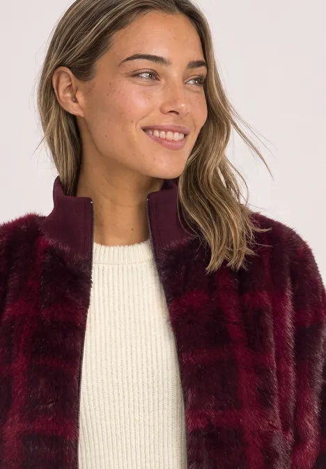 Blouson en fausse fourrure avec des détails en maille et un imprimé plummy brown Eine lächelnde Frau mit weinroter Kunstfelljacke und weißem Pullover, Blick zur Seite.