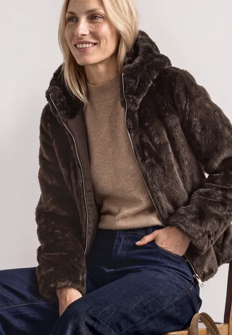 Kurze Fake-Fur-Mix Jacke mit Kapuze auburn brown Frau auf Stuhl, lächelnd in brauner Pelzjacke, beigefarbenem Pullover und blauen Jeans.