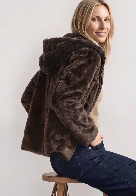 Kurze Fake-Fur-Mix Jacke mit Kapuze auburn brown Frau im braunen, kuscheligen Kunstfellmantel mit Kapuze sitzt seitlich auf einem Hocker und lächelt.