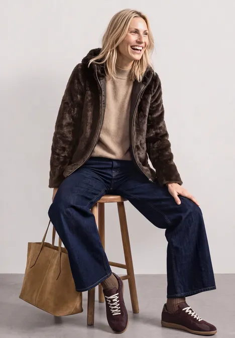 Kurze Fake-Fur-Mix Jacke mit Kapuze auburn brown Frau sitzt lachend auf Hocker, trägt braune Kunstpelzjacke, beigen Pullover, weite Jeans, Sneaker und Tasche.