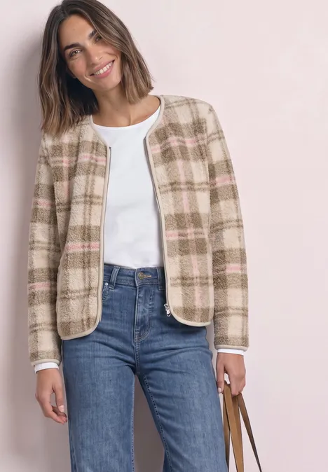 Frau in kariertem Kurzblazer, weißem Top, blauen Jeans und Tasche, lächelt.