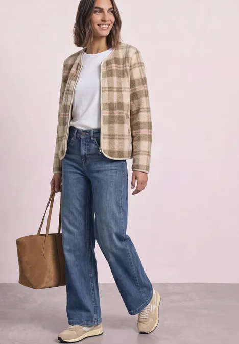 Frau in karierter beige Jacke, weißem T-Shirt, weiten Jeans, beigen Sneakern und brauner Tasche vor hellem Hintergrund.