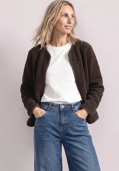 Frau mit brauner Teddyjacke, weißem T-Shirt und blauen Jeans, Hände in den Taschen, seitlicher Blick.