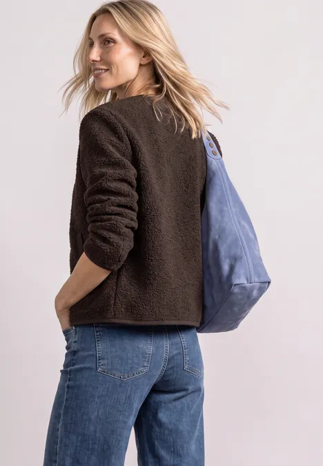 Eine Frau in einer dunkelbraunen Teddyjacke und Jeans trägt eine blaue Tasche und lächelt über die Schulter.